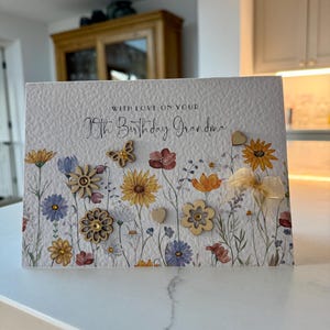 May include: A white card with a floral design and the text "With love on your 90th Birthday Grandma". The card has small wooden flowers and a butterfly attached to it.