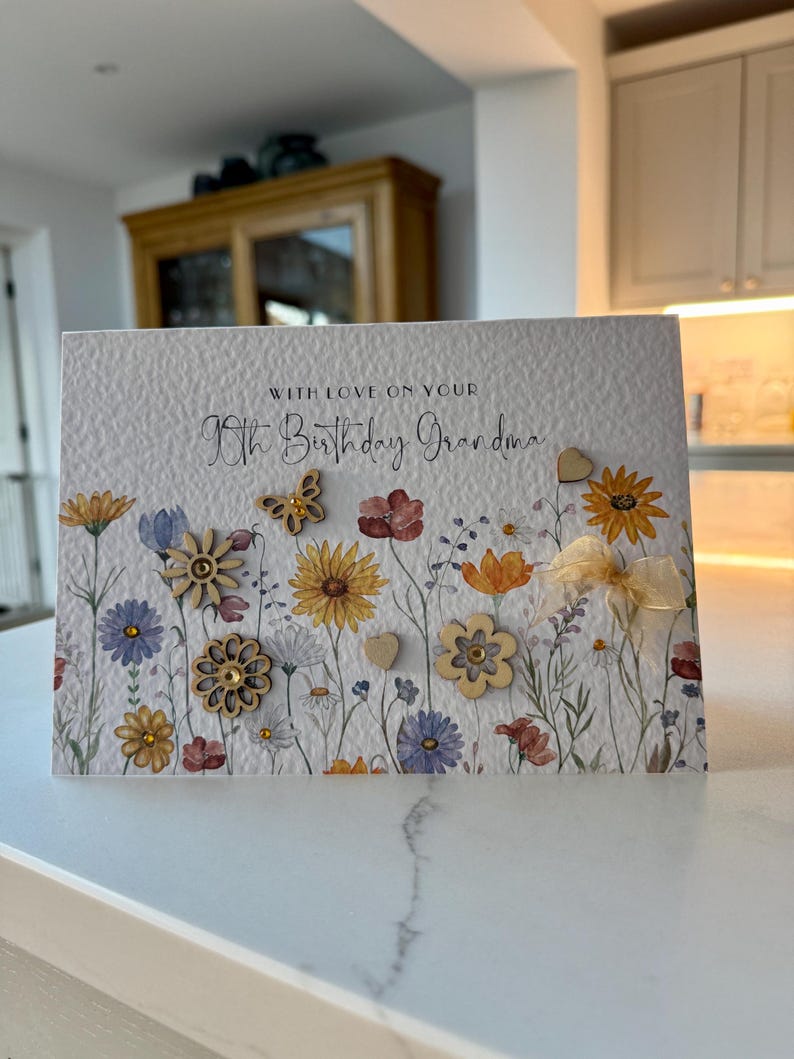 May include: A white card with a floral design and the text "With love on your 90th Birthday Grandma". The card has small wooden flowers and a butterfly attached to it.