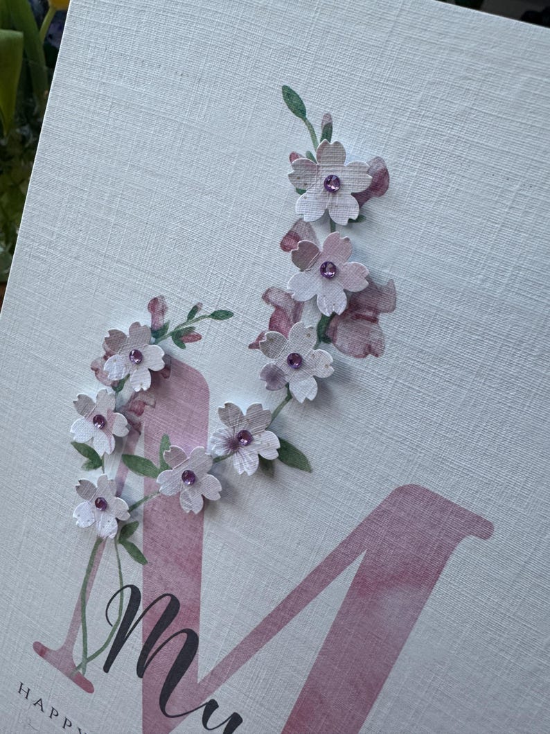 Peut inclure: Carte de v&oelig;ux avec un grand &laquo; M &raquo; rose et le mot &laquo; Happy &raquo; en bas. De d&eacute;licates fleurs en papier avec des centres violets et des feuilles vertes sont dispos&eacute;es le long de la lettre. Le fond est une surface textur&eacute;e blanc cass&eacute;.