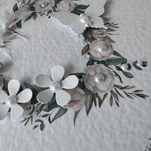 May include: A white greeting card with a floral wreath design. The wreath features paper flowers, a butterfly, and a bow. The card has a textured surface and includes pearl accents.