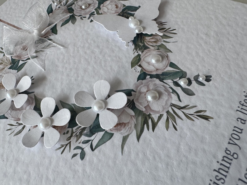 May include: A white greeting card with a floral wreath design. The wreath features paper flowers, a butterfly, and a bow. The card has a textured surface and includes pearl accents.