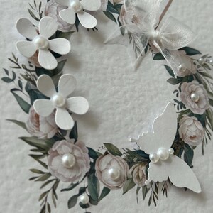 May include: A handcrafted greeting card with a floral wreath design. The wreath incorporates white paper flowers with pearl centres, a white butterfly, and a sheer white bow. The card has a soft, textured background with watercolour-style floral accents.