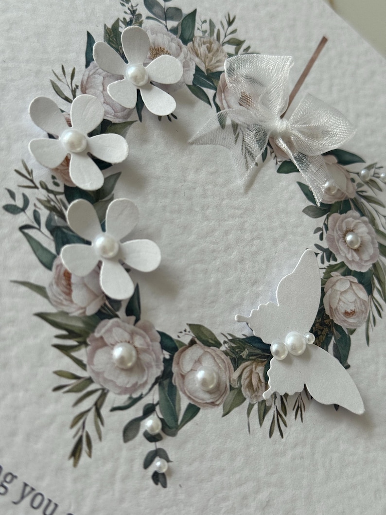 May include: A handcrafted greeting card with a floral wreath design. The wreath incorporates white paper flowers with pearl centres, a white butterfly, and a sheer white bow. The card has a soft, textured background with watercolour-style floral accents.