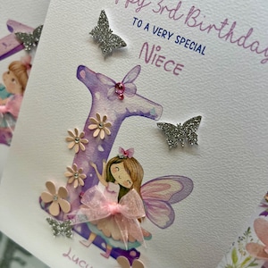 May include: A handmade birthday card with a purple letter "L" decorated with pink flowers and glitter. A fairy with pink wings and a pink bow is standing in front of the letter. The card says "Happy 3rd Birthday to a very special Niece" and is signed "Lucy".