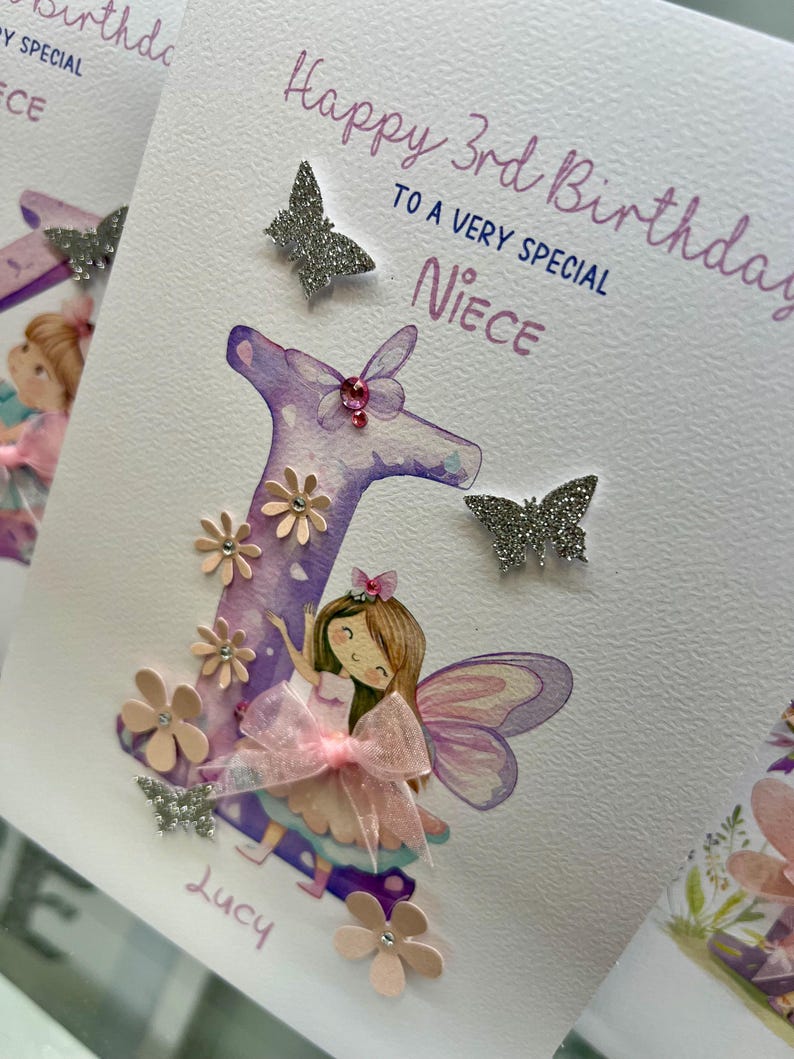May include: A handmade birthday card with a purple letter "L" decorated with pink flowers and glitter. A fairy with pink wings and a pink bow is standing in front of the letter. The card says "Happy 3rd Birthday to a very special Niece" and is signed "Lucy".