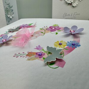 May include: A white card with a pink and purple floral design and the words "Happy Birthday" in black lettering. The card features a pink ribbon, silver butterflies, and purple flowers with pink glitter accents.