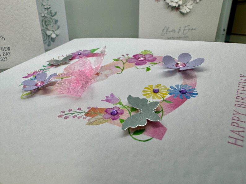 May include: A white card with a pink and purple floral design and the words "Happy Birthday" in black lettering. The card features a pink ribbon, silver butterflies, and purple flowers with pink glitter accents.