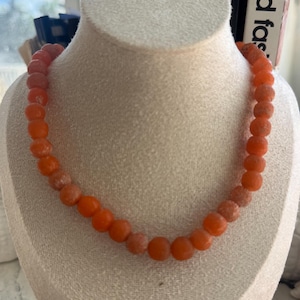 May include: A single-strand necklace composed of round, matte-finish orange beads. The beads exhibit a textured, slightly weathered surface appearance and are strung together in a uniform circular arrangement around a neutral-toned display bust.