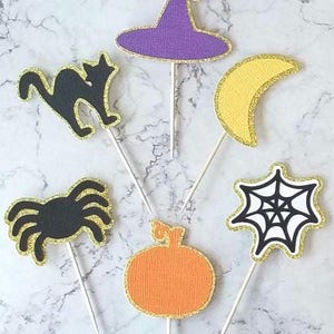 May include: Six Halloween themed cake toppers with gold glitter trim. The toppers feature a witch's hat, a black cat, a crescent moon, a spider, a spider web, and a pumpkin.