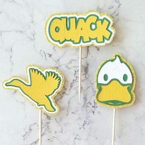 May include: Three yellow duck-themed cupcake toppers. One topper says "QUACK" in yellow letters with a green outline and gold glitter trim. The other two toppers are duck shapes with green and gold glitter trim.