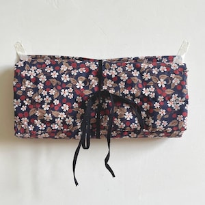 May include: A floral fabric roll-up case with a navy blue background, white flowers, and red berries. The case is tied with a black ribbon and is suspended from the wall by two white clips. The case is likely for storing makeup brushes or art supplies.