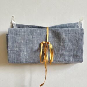 May include: A folded, rectangular, gray linen item is suspended against a white wall, secured with tape. A golden-yellow ribbon is tied around the center, creating a decorative bow. The linen has a subtle, textured appearance.