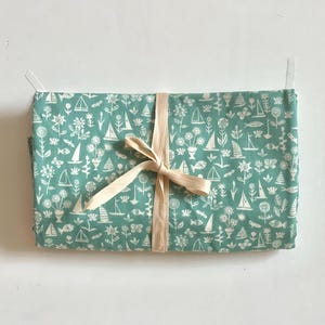 May include: A folded teal fabric with a white nautical pattern of sailboats, flowers, and fish, tied with a cream-colored ribbon. The fabric is rectangular and appears to be a kitchen towel or similar item.