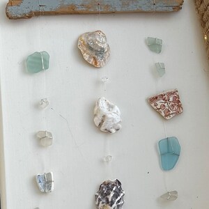May include: A driftwood mobile with seashells and sea glass hanging from it. The mobile is made of a piece of driftwood with string attached to it. The string is holding various seashells and sea glass. The seashells are white and brown. The sea glass is blue and green.