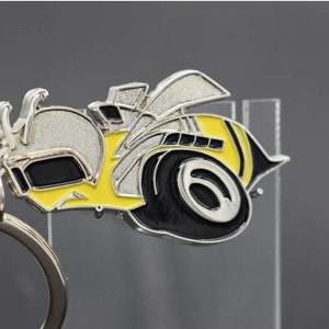Pode incluir: Um chaveiro de metal prateado com um design de abelha amarela e preta. A abelha tem uma asa prateada e um pneu preto com uma borda prateada. O chaveiro tem um anel prateado.