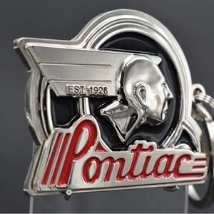Op de afbeelding: Een zilverkleurige metalen sleutelhanger met het Pontiac-logo en een profiel van een indiaans hoofd in reliëf. Het ontwerp bevat de tekst EST. 1926 en het woord Pontiac in rode emaille letters tegen een zwarte achtergrond met zilveren accenten.