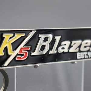 Puede incluir: Un llavero de metal plateado con una etiqueta rectangular negra que dice "K5 Blazer EST. 1969". La "K" es amarilla, el "5" es rojo y el resto del texto es plateado.