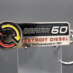 Könnte beinhalten: Ein rechteckiger Metallschlüsselanhänger mit einem kreisförmigen Logo mit roten und gelben Pfeilen auf der linken Seite. Der Text lautet SERIES 60, DETROIT DIESEL und First introduced in 1987. Das Design enthält schwarze, silberne, rote und gelbe Emaille-Akzente.