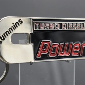 Może przedstawiać: Srebrny i czarny metalowy brelok do kluczy z napisem "Cummins Turbo Diesel Power" w czerwonych i czarnych literach.