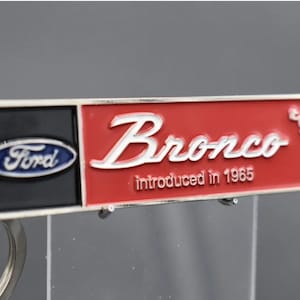 May include: A silver key chain with a red and black enamel rectangular tag. The tag says "Ford" in blue on the black side and "Bronco introduced in 1965" in white on the red side.