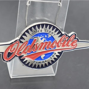 Peut inclure: Une épingle en métal argenté et bleu avec les mots "Oldsmobile" en lettres rouges et blanches. L'épingle présente également un globe avec les mots "Since 1897" et "Final 500" autour.