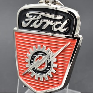 May include: A chrome and enamel Ford emblem keychain. The emblem features the Ford logo in silver on a black background. Below, a red shield with horizontal lines displays a gear and lightning bolt design.