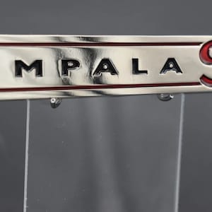Könnte beinhalten: Silberner Schlüsselanhänger mit dem Schriftzug "IMPALA SS" in schwarzer und roter Schrift. Der Schlüsselanhänger hat eine rechteckige Form mit rotem Rand und einem silbernen Schlüsselring.