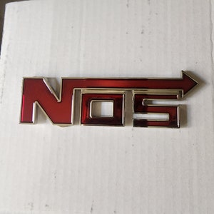 May include: A red and gold metal emblem featuring the letters "NOS" and an arrow. The letters are in a bold, blocky font with a red interior and gold trim. The arrow extends from the "S".