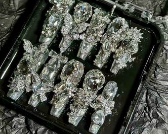 May include: A set of 10 silver nail charms with rhinestones and clear crystals. The charms are arranged in a black tray.