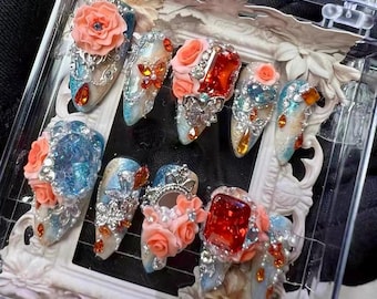May include: A set of artificial nails, each intricately designed. The nails have a base of blue and silver glitter, embellished with rhinestones, peach-coloured roses, and large, colourful gems. They are displayed in a clear case.