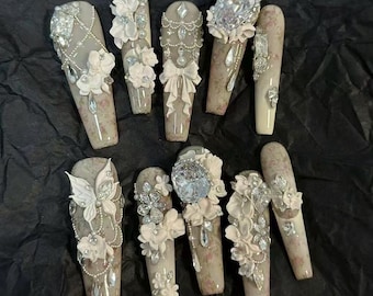 May include: A set of ten long, coffin-shaped artificial nails. Each nail is adorned with white floral details, rhinestones, and pearls. The base colour is a neutral beige with a delicate floral pattern.