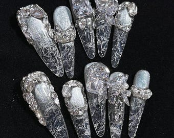 May include: Ten long, clear acrylic nails with silver glitter and crystal detailing. Some nails have large square or teardrop crystals, others feature smaller, clustered gems. The nails are arranged on a dark surface. The nails are approximately 2.5 cm long.