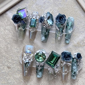 Op de afbeelding: Een set van tien lange, heldere acryl kunstnagels met ingewikkelde ontwerpen. Elke nagel heeft een ander ontwerp, waaronder groene en blauwe edelstenen, zilveren accenten en kleine, donkergroene bloemen.