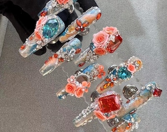 May include: A set of 10 false fingernails with intricate designs. Each nail features a different combination of rhinestones, glitter, and 3D flowers. The flowers are in shades of pink and orange, and the rhinestones are clear, blue, and red.