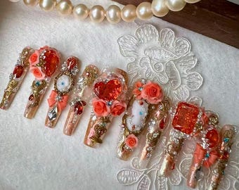 May include: A set of long, embellished artificial nails. Each nail features intricate designs with gold accents, rhinestones, and 3D floral and heart details in shades of red, peach, and white. A pearl necklace is in the background.