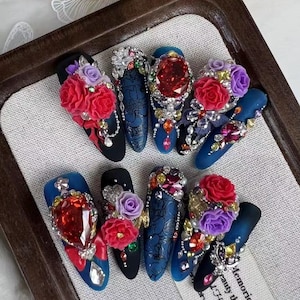 Op de afbeelding: Een assortiment rijkelijk versierde kunstnagels. Elke nagel heeft een zwarte of blauwe basiskleur, versierd met edelstenen, parels en bloemmotieven in rood, paars en wit. Sommige nagels hebben ingewikkelde patronen.