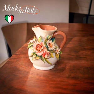 Op de afbeelding: Een crèmekleurige keramische kan met een bruin handvat en rand, versierd met gebeeldhouwde perzikrozen en witte madeliefjes. De kan is versierd met groene bladeren en de tekst "Made in Italy" is zichtbaar.