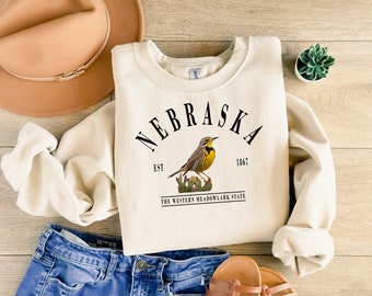 May include: A cream-colored sweatshirt with the text "NEBRASKA EST 1867 THE WESTERN MEADOWLARK STATE" and a yellow bird illustration.