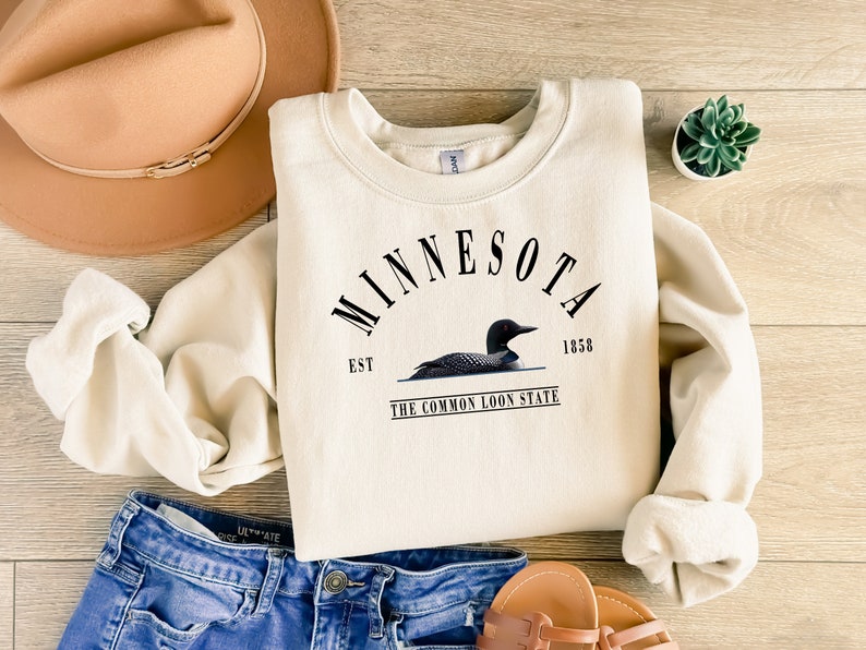 May include: A cream-colored sweatshirt with a black graphic print of a loon on a lake. The graphic includes the text "MINNESOTA EST 1858 THE COMMON LOON STATE".