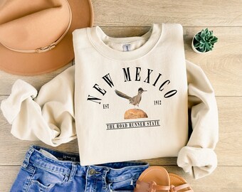 May include: A cream-colored sweatshirt with a graphic design featuring a roadrunner bird and the text "New Mexico Est. 1912 The Road Runner State".