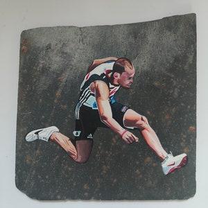 May include: A painting on a slate tile of an athlete in mid-air, wearing a white, black, and red uniform, with a red wristband, and white shoes. The athlete is jumping over a hurdle.