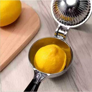 May include: A stainless steel lemon squeezer with a yellow lemon inside. The squeezer is open, ready to be pressed. A wooden cutting board and another lemon are in the background.