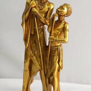May include: Gold-toned statue of a couple standing side-by-side. The man is holding a staff and the woman is holding a smaller staff. Both figures are wearing traditional clothing and jewellery.