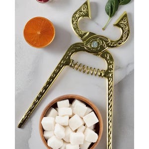 Puede incluir: Una pinza de azúcar dorada con detalles ornamentados, colocada sobre un pequeño cuenco de madera lleno de cubos de azúcar blancos. La imagen también incluye una rodaja de naranja, un cuenco pequeño de bayas rojas y hojas verdes.
