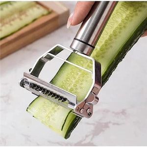 May include: A stainless steel vegetable peeler is shown in use, removing a strip of green cucumber. The peeler has a silver handle and a blade with small teeth. A wooden cutting board with cucumber slices is in the background.