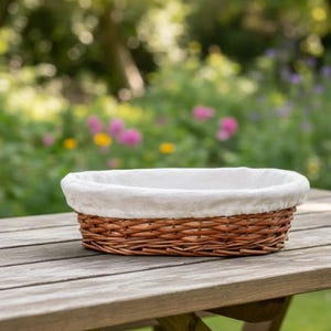 Op de afbeelding: Een bruine, ovale rieten mand met een witte stoffen voering staat op een houten picknicktafel. De mand is leeg. De achtergrond toont een vage tuin met groene bladeren en kleurrijke bloemen.