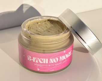 May include: A small, clear jar with a silver lid, containing a light brown cream. The jar has a pink label with the text "B-ITCH NO MORE. MOSQUITO BITE RELIEF CLAY 1 OZ."