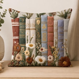 May include: A square throw pillow featuring an embroidered design of a row of books in shades of red, green, white, blue, and tan. The spines are decorated with intricate floral embroidery, including daisies and other small blossoms in various colors.