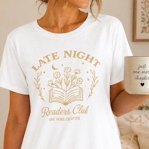 May include: A white short-sleeve t-shirt dress featuring a gold graphic print that reads Late Night Readers Club One More Chapter, accompanied by illustrations of an open book, flowers, stars, and a crescent moon, paired with a white ceramic mug.