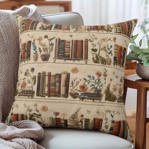 May include: A decorative throw pillow with a cream-colored background. The pillow features a repeating pattern of bookshelves, potted plants, and flowers in shades of brown, orange, and green. The pillow is square and appears to be made of a soft fabric.